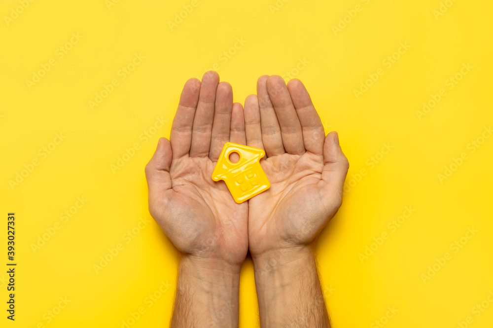 House symbol in male hands on yellow background flat lay top view. Model of plastic yellow house. Concept of buying home, apartment. Renting a house, real estate. Mortgage, real estate insurance House symbol in male hands on yellow background flat lay top view. Model of plastic yellow house. Concept of buying home, apartment. Renting a house, real estate. Mortgage, real estate insurance