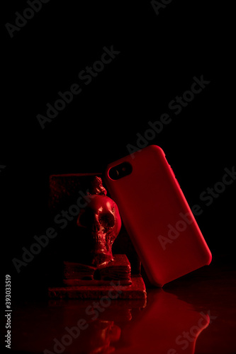 Telefono móvil con funda de color rojo apoyado en una calavera con un fondo negro y luces rojas.
 photo