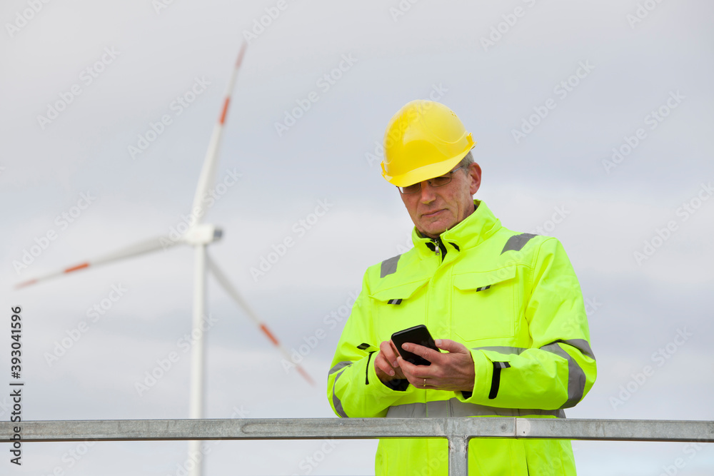 Mature engineer with protective clothing looking at a smart phone in front of a wind turbine Mature engineer with protective clothing looking at a smart phone in front of a wind turbine