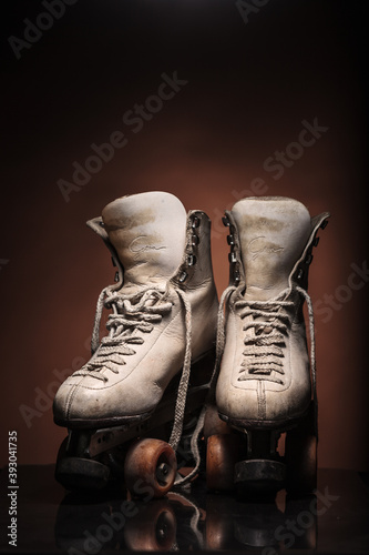 Old heavily used worn-out pair of white roller skates showing their boot tongues with some dirt and untied shoelace on reflective surface and warm brown background with upper space for titles