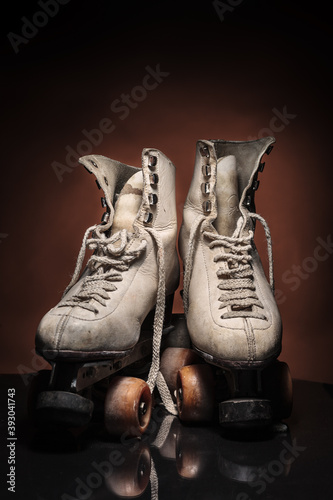 Old heavily used worn-out pair of white roller skates with some dirt and untied detailed shoelace buckles on reflective surface and warm brown background