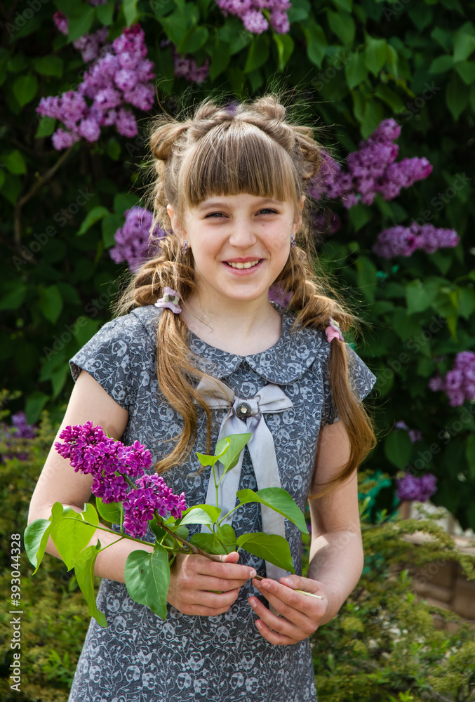 A little girl with beautiful braids stands by a blooming tree, holding lilac flowers in her hands A little girl with beautiful braids stands by a blooming tree, holding lilac flowers in her hands