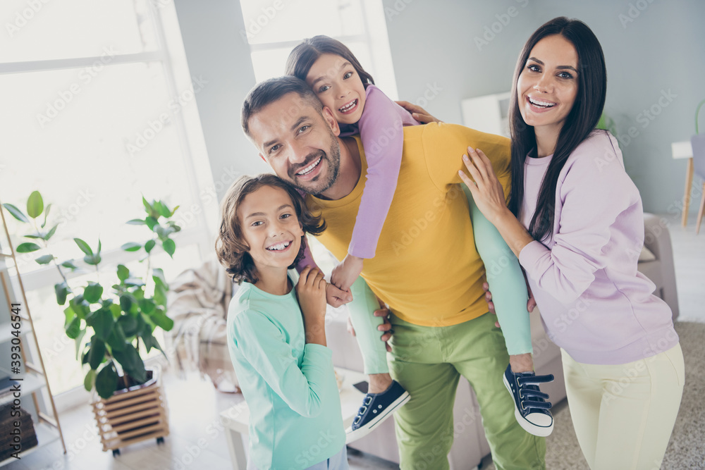 Photo of full big family four people daddy hold piggy back daughter cuddle relatives wear colorful sweater pants in living room indoors