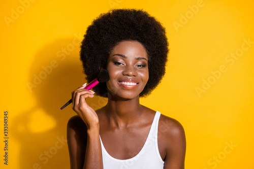 Close-up portrait of attractive cheery wavy-haired girl applying bronzer on cheek isolated on bright yellow color background