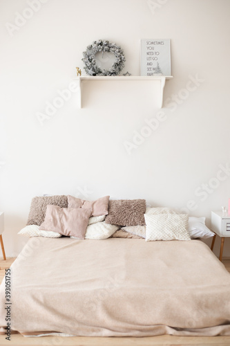 a cozy bedroom in beige colors, a bed with a blanket and pillows, bedside tables, a white wall, a white wooden shelf with a Christmas wreath and a photo frame with the inscription