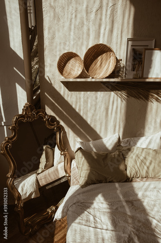Modern boho style home interior design. Bohemian bedroom with bed, pillows, straw and rattan decorations, vintage retro mirror, palm leaf, concrete wall. Sunlight shadows on the wall. Hygge concept.