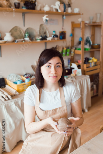 Young beautiful woman sitting at her workspace in ceramics studio with wet clay for modeling