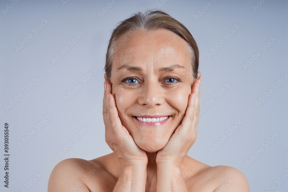 Portrait of a smiling mature woman with her hands to her cheeks, looking at the camera against a gray background. Anti-wrinkle facial skin care concept. Portrait of a smiling mature woman with her hands to her cheeks, looking at the camera against a gray background. Anti-wrinkle facial skin care concept.