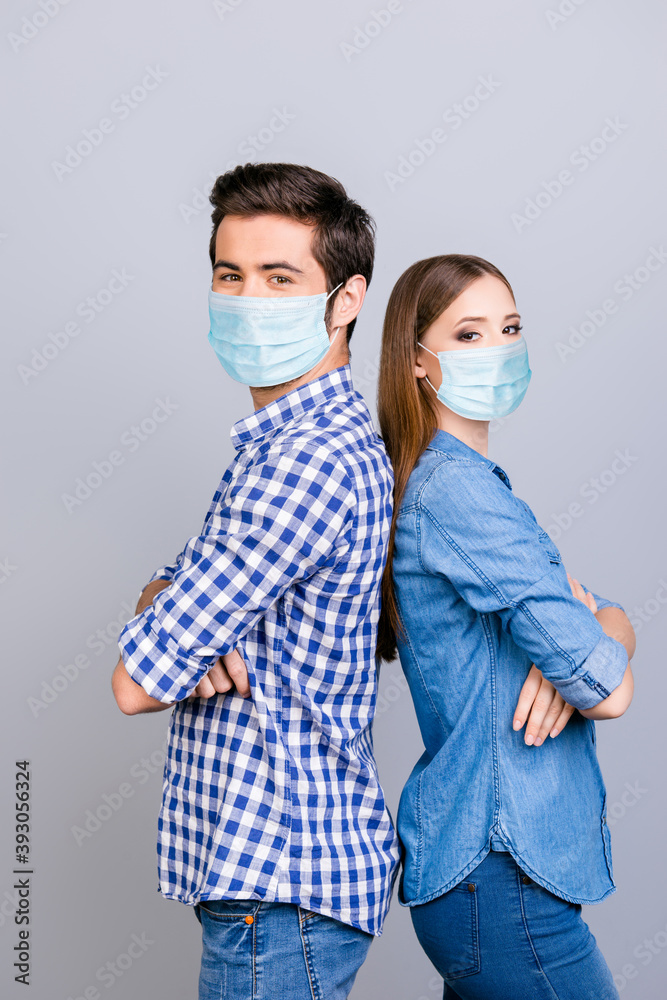 Vertical photo portrait of two young lovers looking at the camera standing back to back wearing blue face masks with crossed hands isolated on white colored background