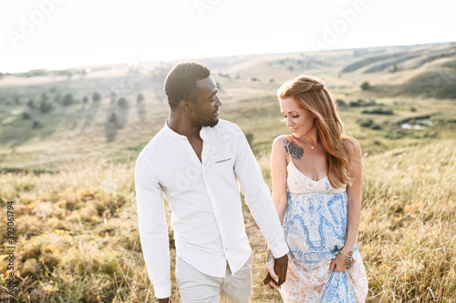 Romantic couple in love walking on the meadow. A beautiful pregnant red haired wife and her handsome black husband hugging outdoors