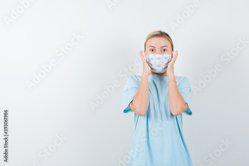  young lady holding hands near face in t-shirt, mask and looking perplexed. front view. © ABUATOP