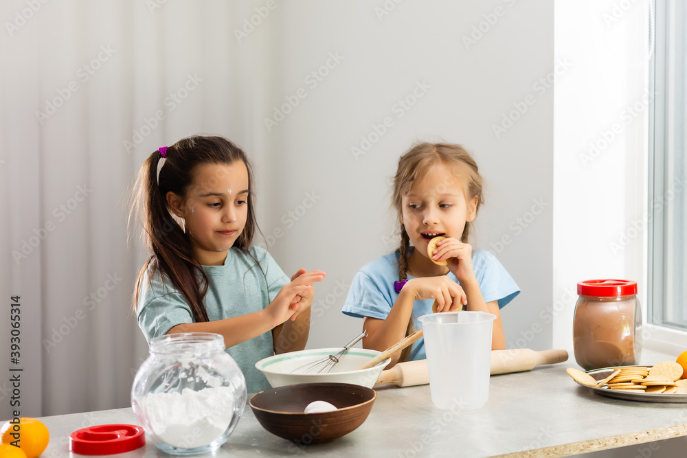 Two little girls in the kitchen prepare food, a dessert for the family. As they learn to cook they start playing with flour and smiling each other. Concept of: cooking classes, family, education. Two little girls in the kitchen prepare food, a dessert for the family. As they learn to cook they start playing with flour and smiling each other. Concept of: cooking classes, family, education.