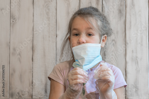 Little girl with medical mask and disposable gloves checks gloves for integrity, anti-coronavirus in pink t-shirt, wooden background photos