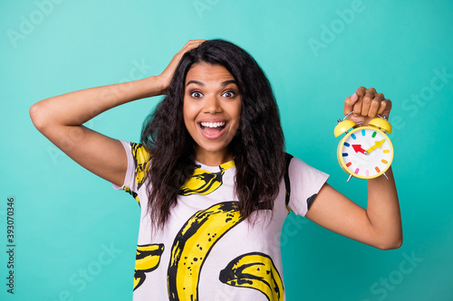 Photo of young girl hold alarm clock hand head open mouth wear banana print t-shirt isolated teal color background
