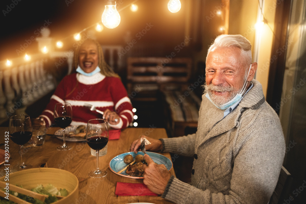Multiracial senior people celebrate christmas together with dinner outdoor while wearing safety mask for coronavirus outbreak - Soft focus on man face