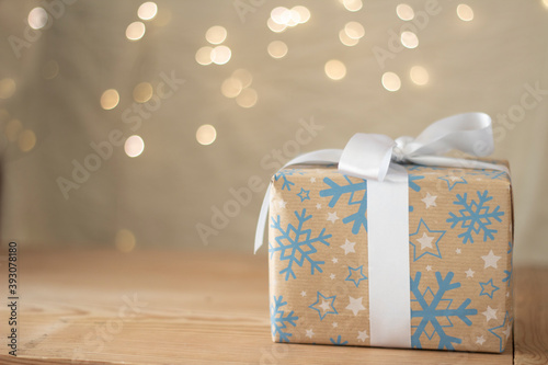 christmas gift wrapped in recycled paper with blue flakes and white ribbon background of unfocused white christmas lights. Concept of celebrating Christmas-giving. photo