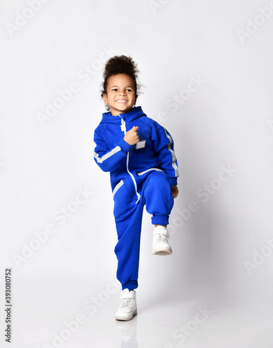 Happy sporty dark-skinned kid girl in blue trendy jumpsuit in motion does fitness exercises, warmup, marching, holding foot up, stands on one leg in sports school