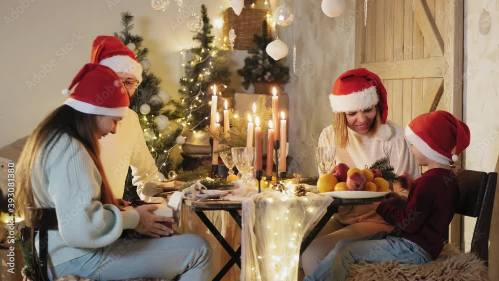 Family of four at the dinner table at Christmas. Everyone in Santa hats, dad and mom give gifts to children Family of four at the dinner table at Christmas. Everyone in Santa hats, dad and mom give gifts to children
