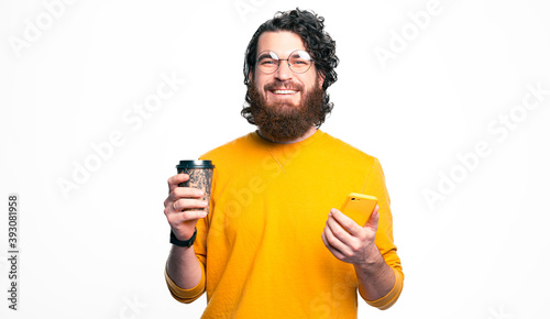 Happy bearded man wearing round glasses and looking confident at the camera and holding coffee and smartphone.