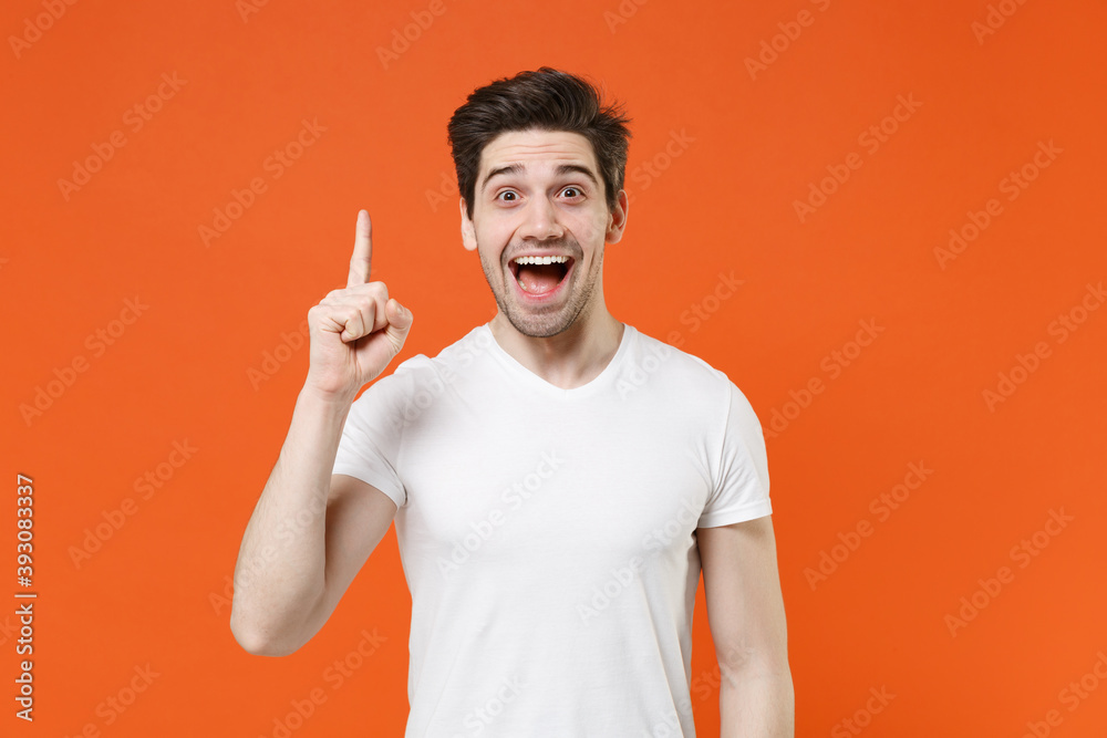 Excited young man 20s in basic casual white blank empty t-shirt standing holding index finger up with great new idea looking camera isolated on bright orange colour wall background, studio portrait. Excited young man 20s in basic casual white blank empty t-shirt standing holding index finger up with great new idea looking camera isolated on bright orange colour wall background, studio portrait.