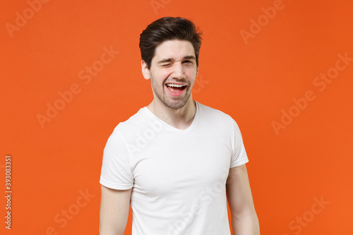 Blinking cheerful funny handsome attractive young man 20s wearing basic casual white blank empty t-shirt standing looking camera isolated on bright orange colour wall background, studio portrait. © ViDi Studio