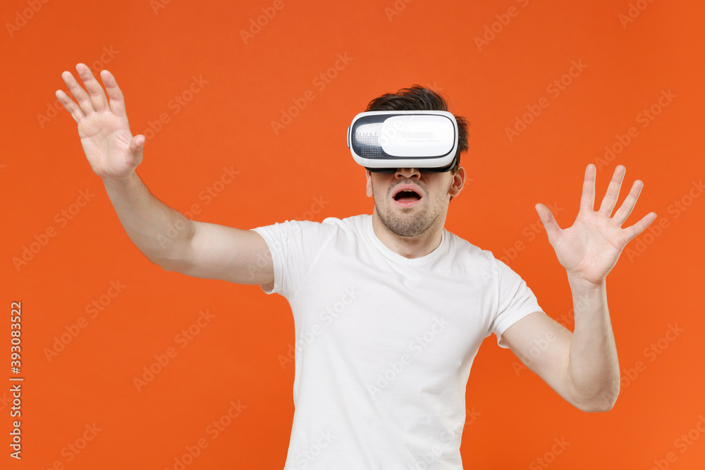 Shocked amazed young man 20s in basic casual white blank empty t-shirt standing watching in vr headset gadget touch something like push click on button isolated on orange background studio portrait. Shocked amazed young man 20s in basic casual white blank empty t-shirt standing watching in vr headset gadget touch something like push click on button isolated on orange background studio portrait.