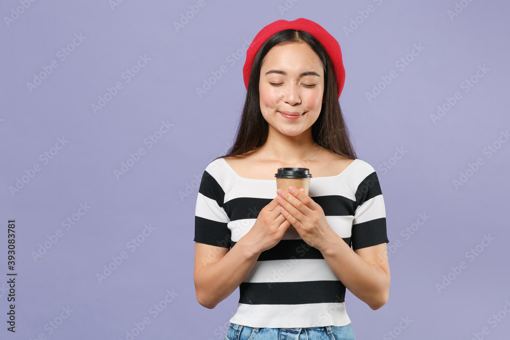 Smiling pretty young brunette asian woman 20s in striped t-shirt red beret standing holding paper cup of coffee or tea keeping eyes closed isolated on pastel violet colour background studio portrait. Smiling pretty young brunette asian woman 20s in striped t-shirt red beret standing holding paper cup of coffee or tea keeping eyes closed isolated on pastel violet colour background studio portrait.