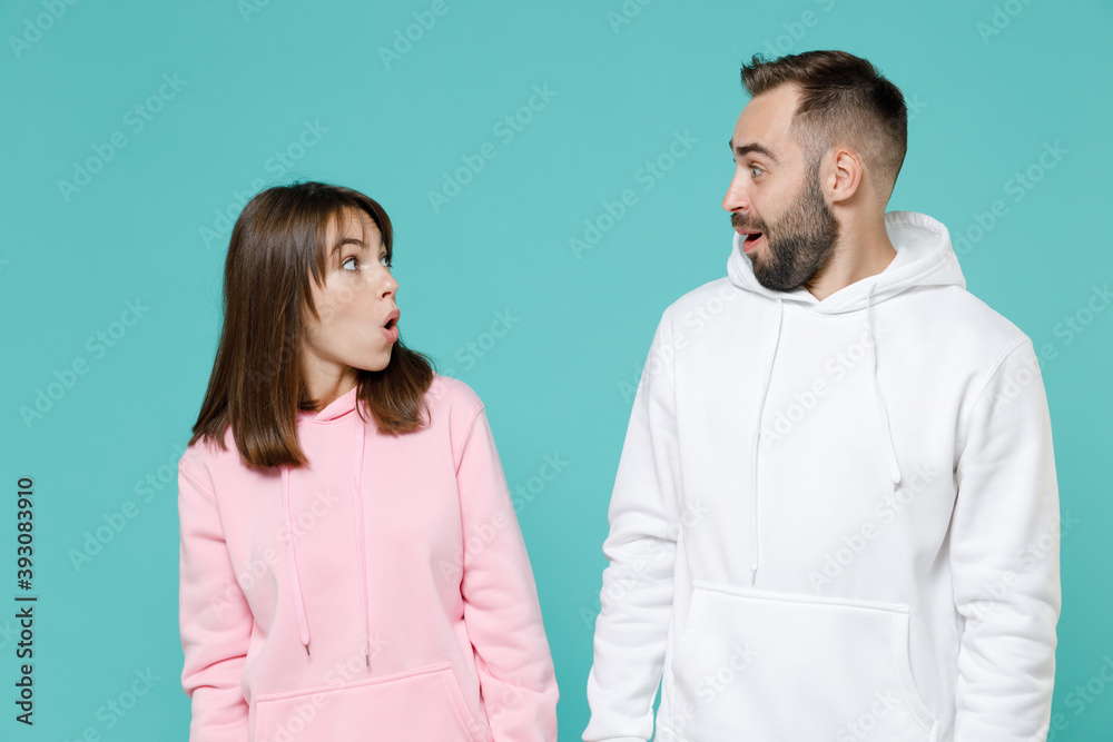 Shocked amazed young couple two friends man woman 20s in white pink casual hoodie standing keeping mouth open looking at each other isolated on blue turquoise colour wall background studio portrait. Shocked amazed young couple two friends man woman 20s in white pink casual hoodie standing keeping mouth open looking at each other isolated on blue turquoise colour wall background studio portrait.