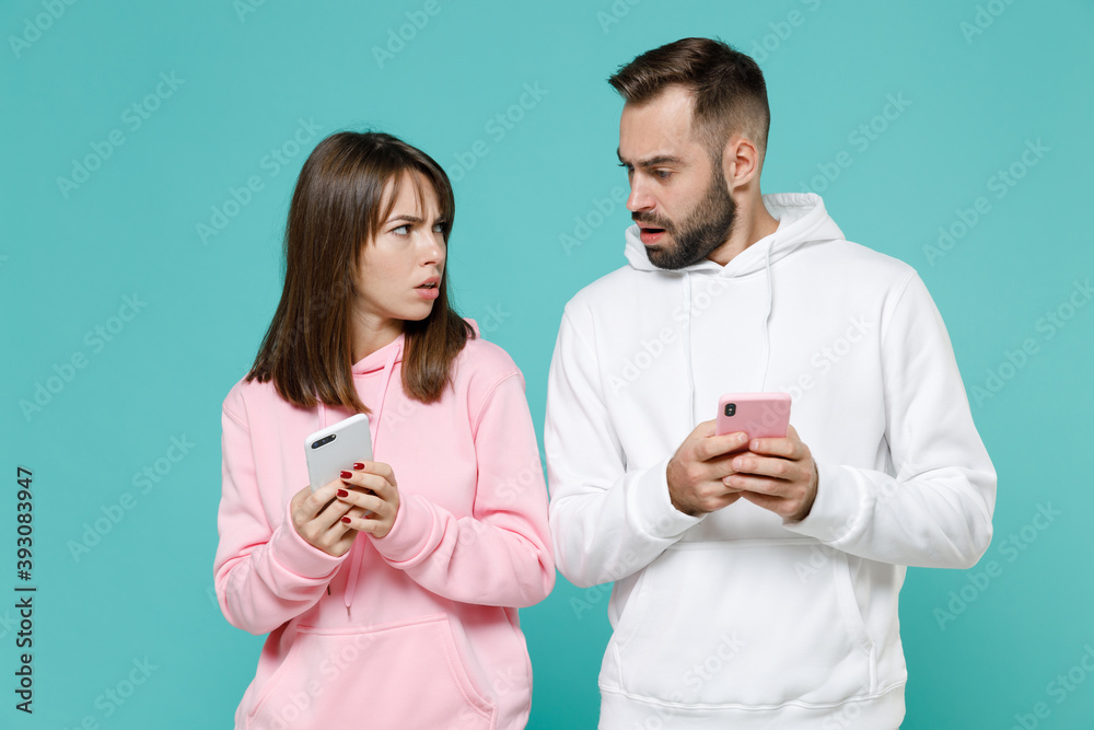 Displeased worried young couple two friends man woman 20s wearing white pink casual hoodie using mobile cell phone typing sms message isolated on blue turquoise colour wall background studio portrait. Displeased worried young couple two friends man woman 20s wearing white pink casual hoodie using mobile cell phone typing sms message isolated on blue turquoise colour wall background studio portrait.
