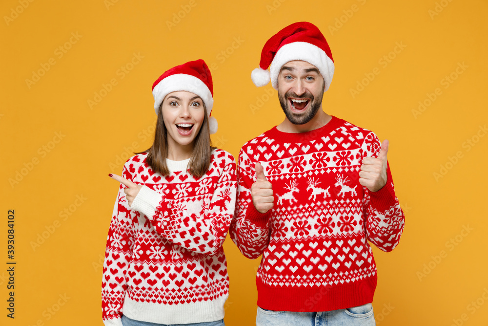 Excited young Santa couple friends man woman in red sweater Christmas hat pointing index finger aside showing thumbs up isolated on yellow background. Happy New Year celebration merry holiday concept. Excited young Santa couple friends man woman in red sweater Christmas hat pointing index finger aside showing thumbs up isolated on yellow background. Happy New Year celebration merry holiday concept.