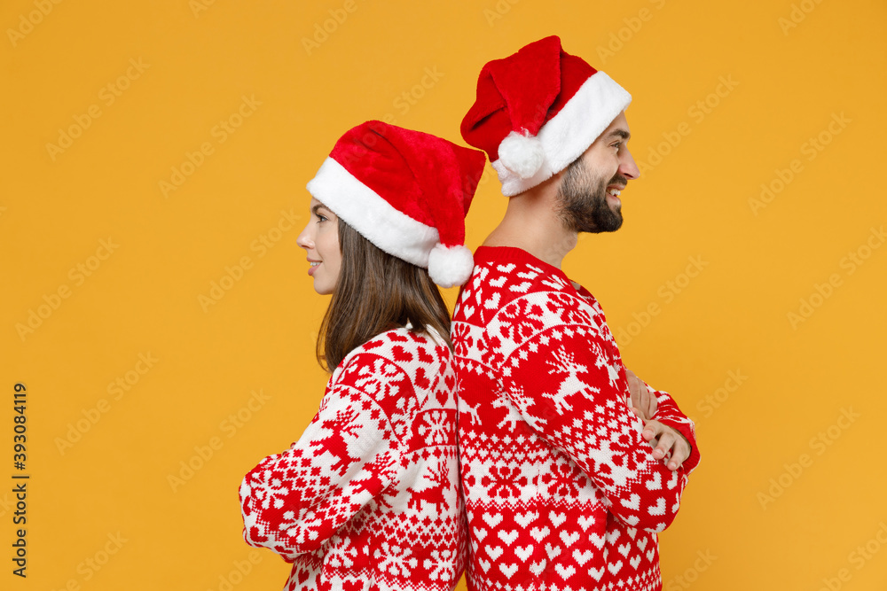 Side view of smiling young Santa couple friends man woman 20s in red sweater Christmas hat holding hands crossed isolated on yellow background studio. Happy New Year celebration merry holiday concept. Side view of smiling young Santa couple friends man woman 20s in red sweater Christmas hat holding hands crossed isolated on yellow background studio. Happy New Year celebration merry holiday concept.