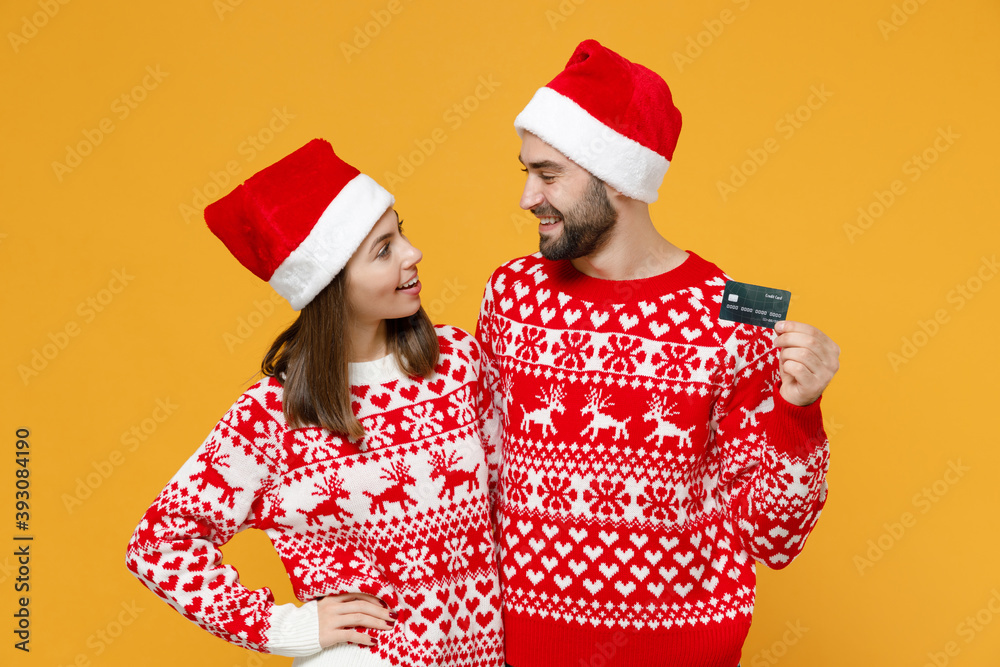 Smiling young Santa couple friends man woman wearing red sweater Christmas hat hold credit bank card isolated on yellow background studio portrait. Happy New Year celebration merry holiday concept.