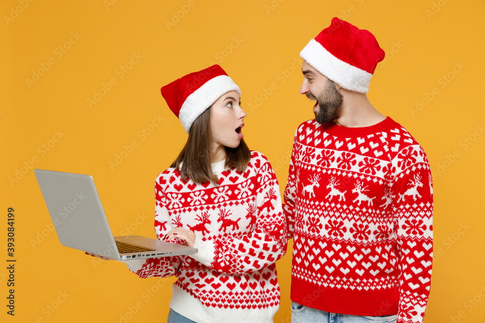 Shocked young Santa couple friends man woman in red sweater Christmas hat working on laptop pc computer isolated on yellow background studio portrait. Happy New Year celebration merry holiday concept. Shocked young Santa couple friends man woman in red sweater Christmas hat working on laptop pc computer isolated on yellow background studio portrait. Happy New Year celebration merry holiday concept.