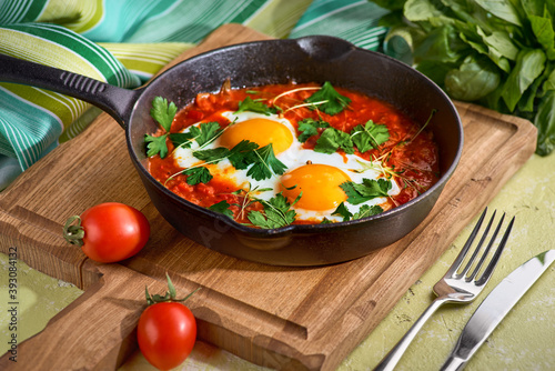 delicious middle east shakshuka - fried eggs, onion, bell pepper, tomatoes, chilli and spices in cast iron stewpan with kitchen towel on old background, authentic recipe