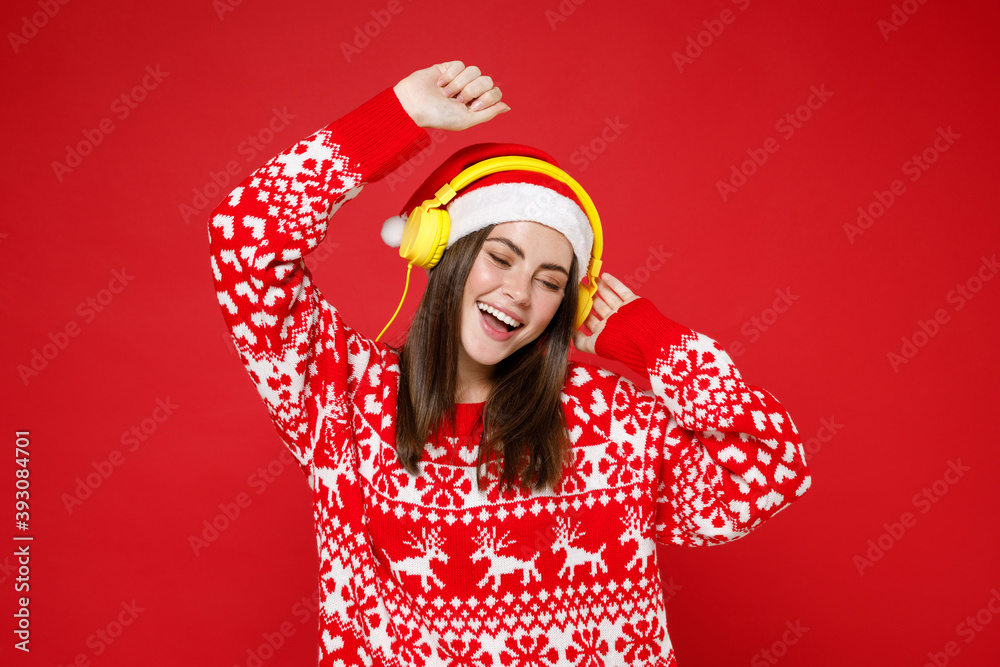 Funny young Santa woman 20s in sweater Christmas hat listening music with headphones dancing rising hands isolated on red background studio portrait. Happy New Year celebration merry holiday concept. Funny young Santa woman 20s in sweater Christmas hat listening music with headphones dancing rising hands isolated on red background studio portrait. Happy New Year celebration merry holiday concept.