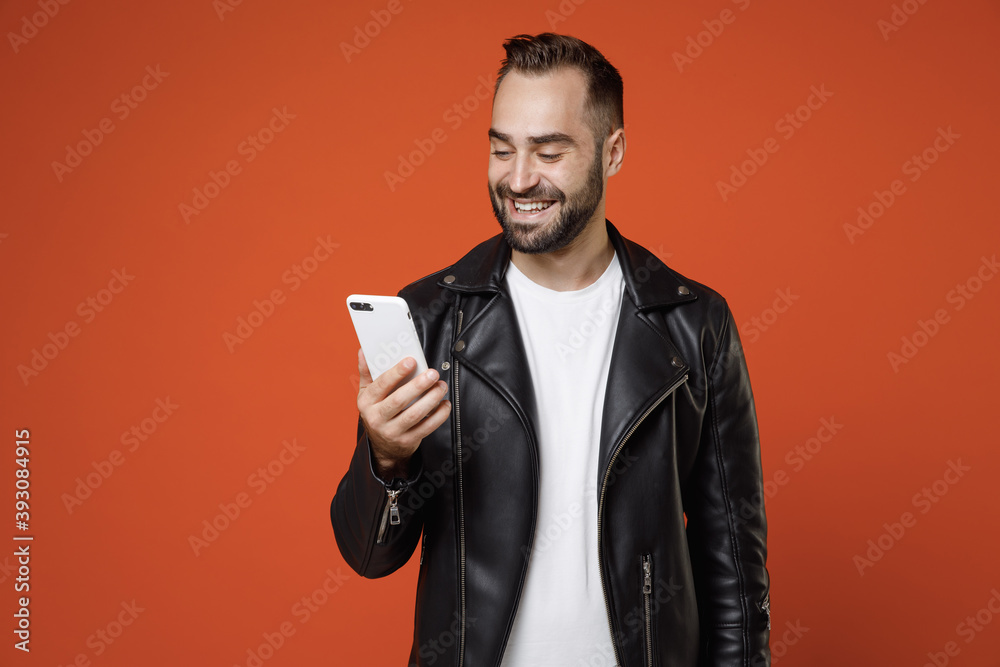 Smiling funny young bearded man 20s wearing basic white t-shirt black leather jacket standing using mobile cell phone typing sms message isolated on bright orange colour background studio portrait. Smiling funny young bearded man 20s wearing basic white t-shirt black leather jacket standing using mobile cell phone typing sms message isolated on bright orange colour background studio portrait.