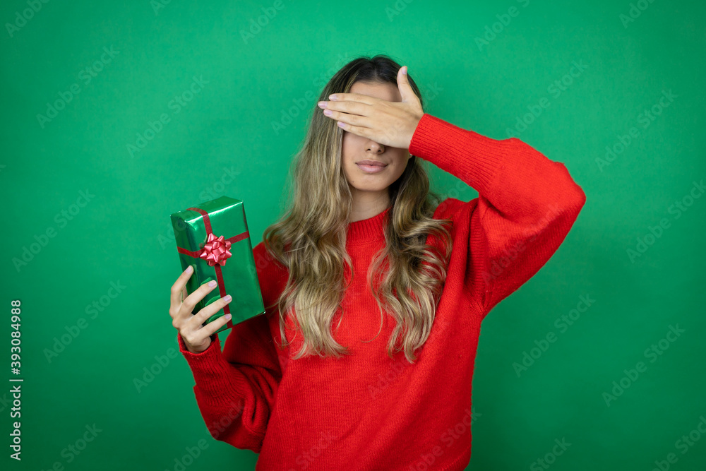 Young beautiful girl holding gift over isolated green background serious and covering her eyes with her hand Young beautiful girl holding gift over isolated green background serious and covering her eyes with her hand