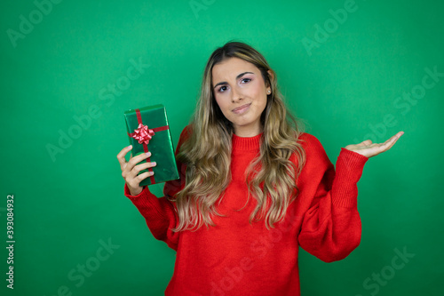 Young beautiful girl holding gift over isolated green background clueless and confused expression with arms and hands raised