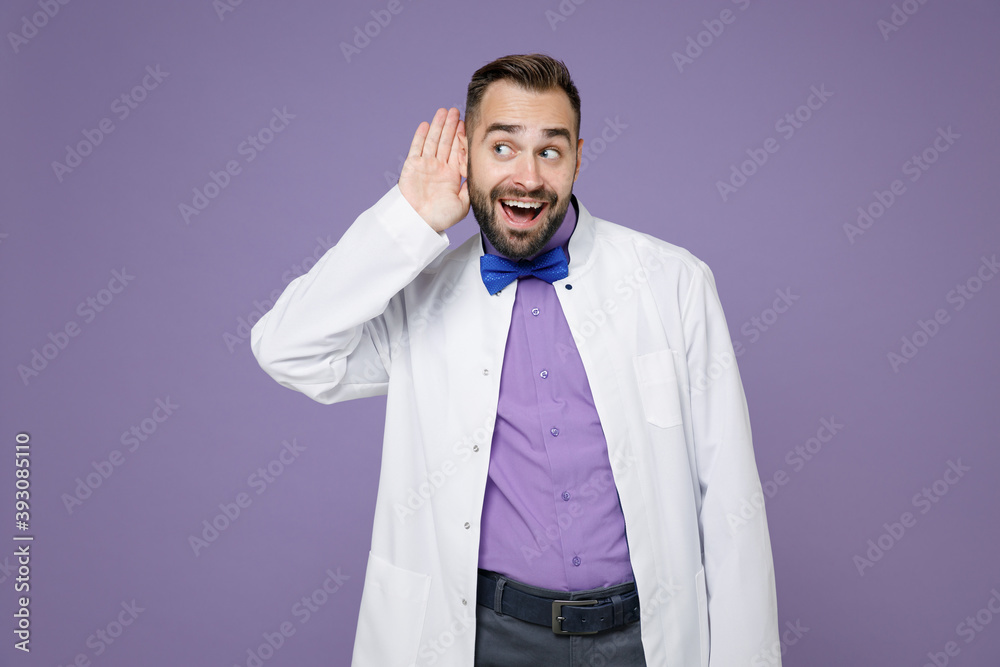 Curious excited young bearded doctor man in white medical gown try to hear you overhear listening intently isolated on violet background studio portrait. Healthcare personnel health medicine concept. Curious excited young bearded doctor man in white medical gown try to hear you overhear listening intently isolated on violet background studio portrait. Healthcare personnel health medicine concept.