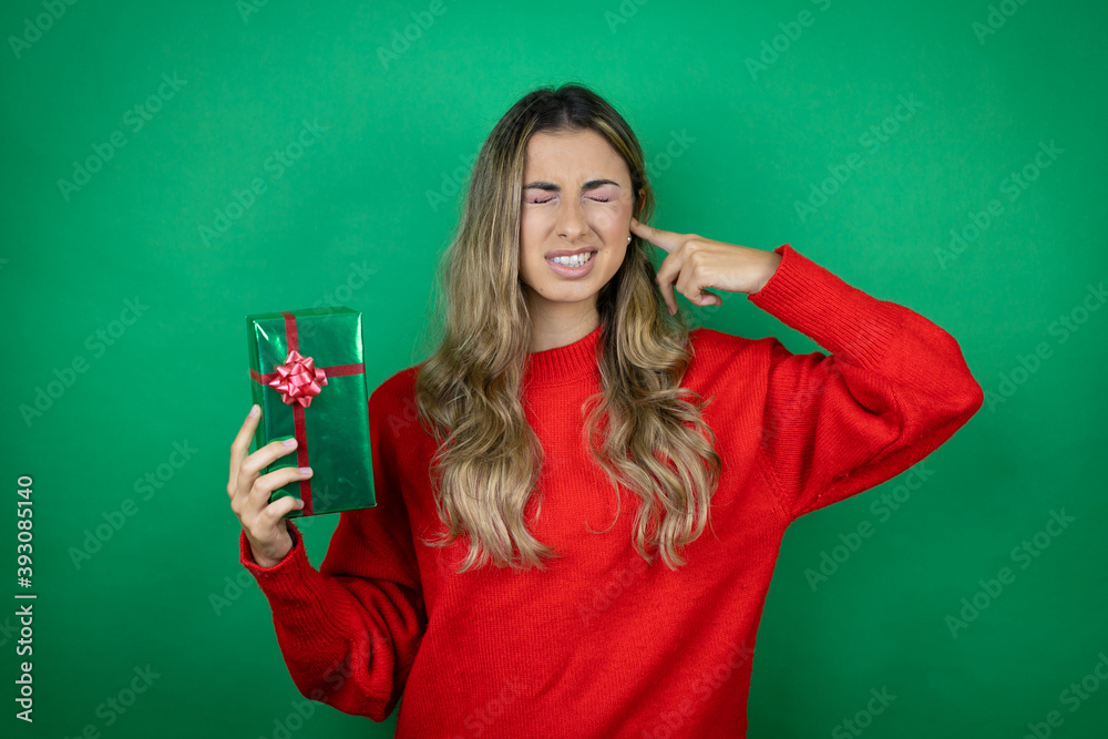 Young beautiful girl holding gift over isolated green background covering ears with fingers with annoyed expression for the noise of loud music. Deaf concept. Young beautiful girl holding gift over isolated green background covering ears with fingers with annoyed expression for the noise of loud music. Deaf concept.