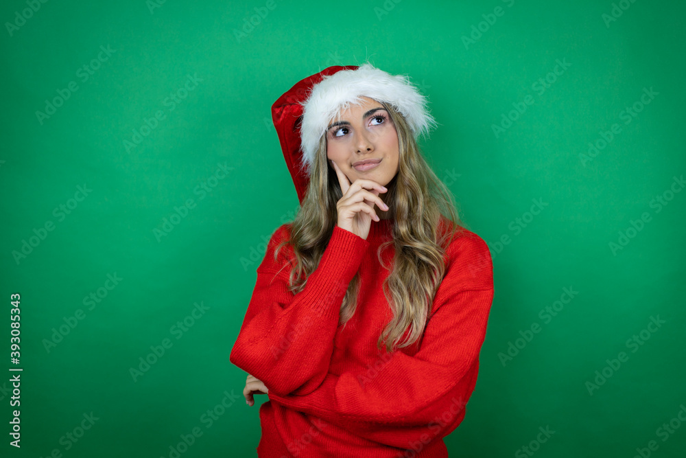 Young beautiful girl wearing Christmas Santa hat holding gift over isolated green background thinking and looking to the side Young beautiful girl wearing Christmas Santa hat holding gift over isolated green background thinking and looking to the side