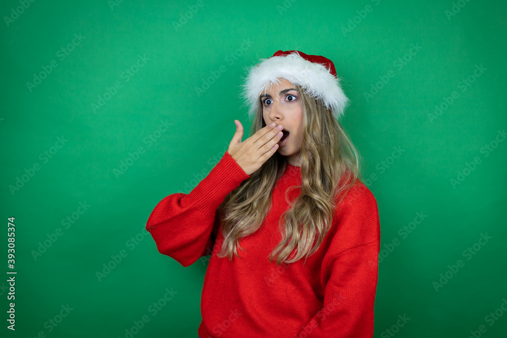 Young beautiful girl wearing Christmas Santa hat holding gift over isolated green background with her hand over her mouth and surprised, looking side Young beautiful girl wearing Christmas Santa hat holding gift over isolated green background with her hand over her mouth and surprised, looking side