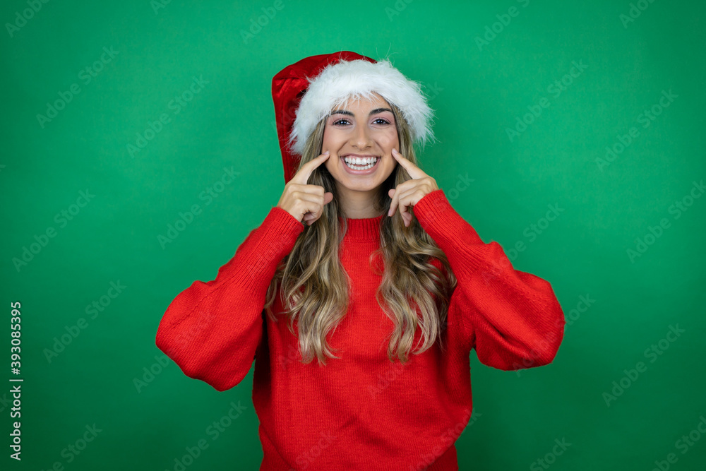 Young beautiful girl wearing Christmas Santa hat holding gift over isolated green background smiling confident showing and pointing with fingers teeth and mouth Young beautiful girl wearing Christmas Santa hat holding gift over isolated green background smiling confident showing and pointing with fingers teeth and mouth