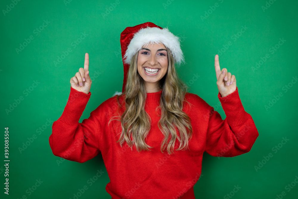 Young beautiful girl wearing Christmas Santa hat holding gift over isolated green background Young beautiful girl wearing Christmas Santa hat holding gift over isolated green background