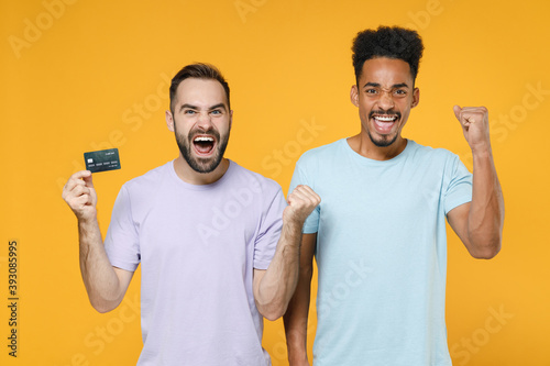 Happy young two friends european african american men 20s in violet blue casual t-shirts hold credit bank card doing winner gesture isolated on bright yellow colour wall background studio portrait.