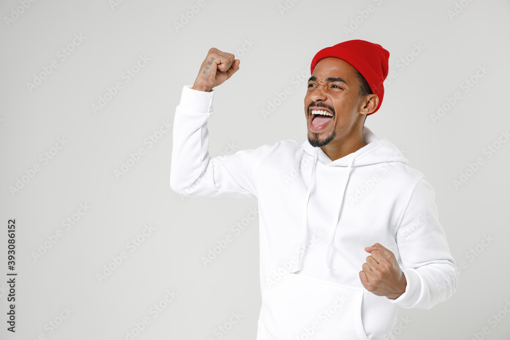 Happy joyful young african american man 20s years old in basic streetwear hoodie standing doing winner gesture clenching fists looking aside isolated on white colour wall background studio portrait. Happy joyful young african american man 20s years old in basic streetwear hoodie standing doing winner gesture clenching fists looking aside isolated on white colour wall background studio portrait.
