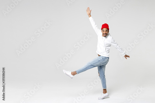 Full length side view of excited funny young african american man 20s in casual streetwear hoodie dancing rising leg spreading hands looking camera isolated on white wall background, studio portrait.