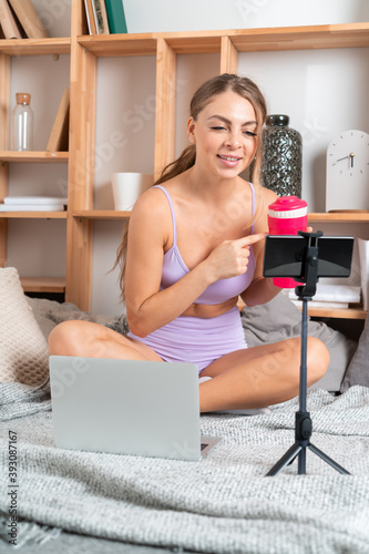 Beautiful young fit woman in pink sportswear making video about sports nutritional supplements for social network with smartphone. Online fitness and vlogging concept.