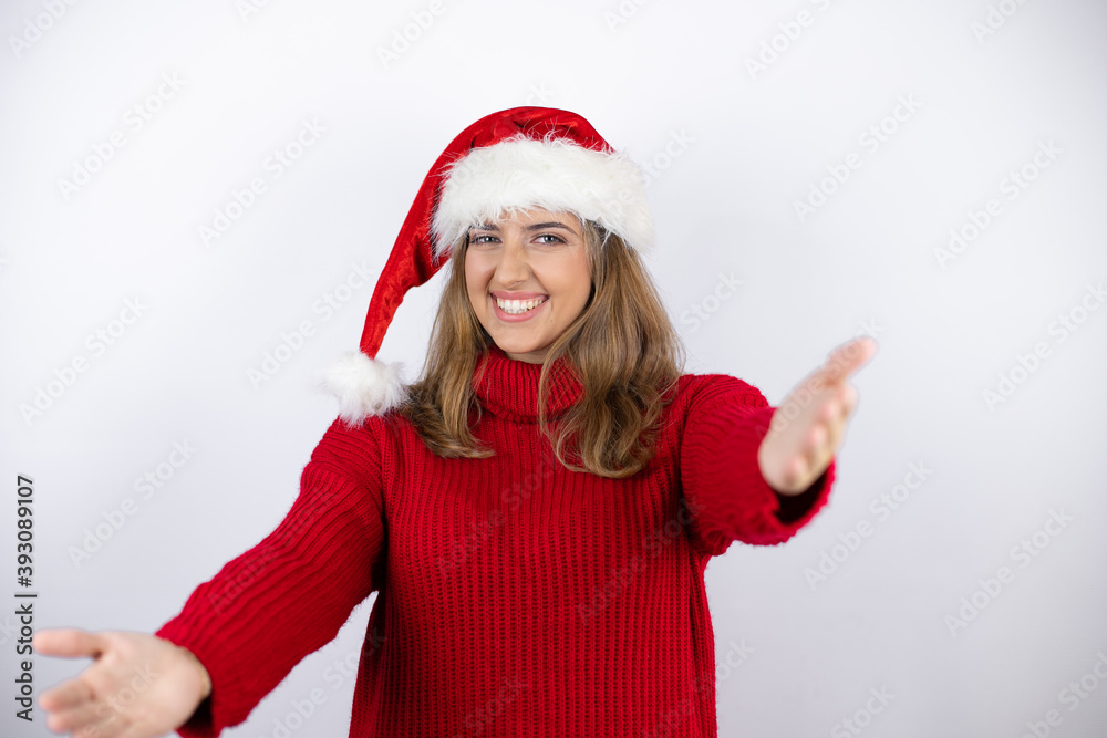 Young pretty blonde woman wearing a red casual sweater and a christmas hat over white background looking at the camera smiling with open arms for hug Young pretty blonde woman wearing a red casual sweater and a christmas hat over white background looking at the camera smiling with open arms for hug
