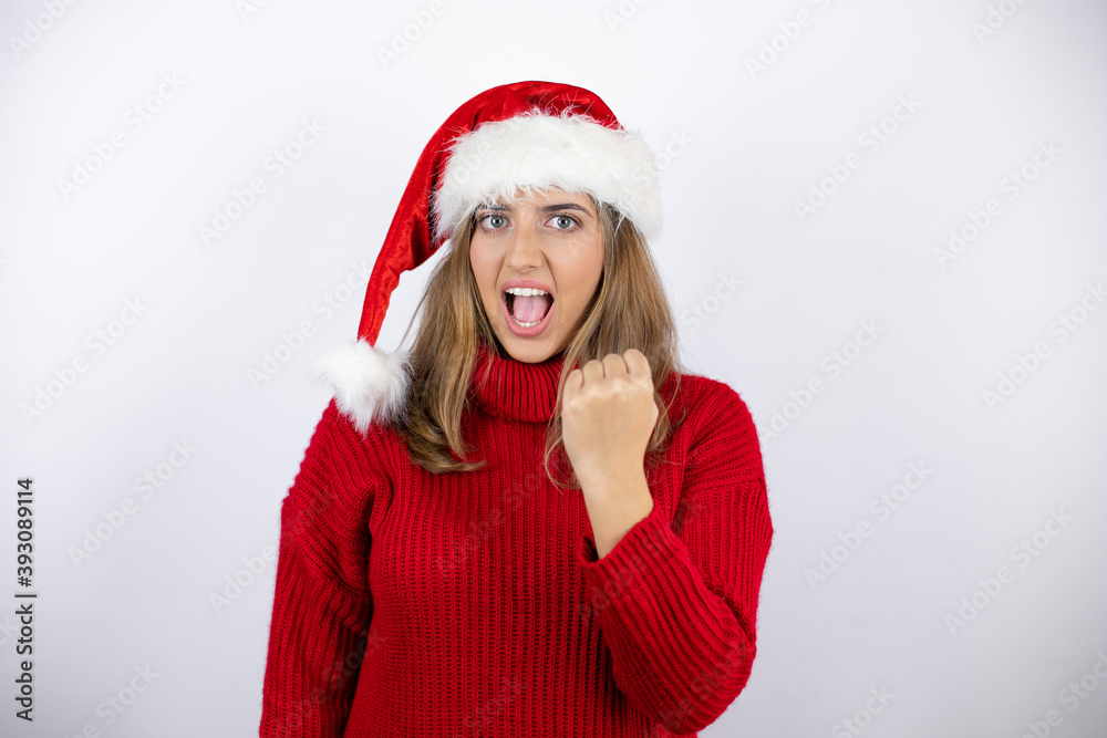 Young blonde woman wearing a red casual sweater and a christmas hat over white background angry and mad raising fist frustrated and furious while shouting with anger. Rage and aggressive concept. Young blonde woman wearing a red casual sweater and a christmas hat over white background angry and mad raising fist frustrated and furious while shouting with anger. Rage and aggressive concept.